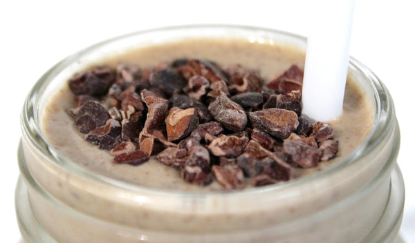 A close-up of Vitasave's Superfood Smoothie "Maca Monkey" in a clear glass, showcasing its creamy texture and rich, earthy color with fresh banana slices and cacao nibs on top.