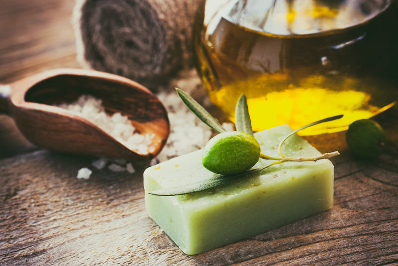 A neatly arranged display of five all-natural bathroom essentials, including eco-friendly soap bars, bamboo toothbrushes, reusable cotton pads, natural loofahs, and herbal bath salts, set on a wooden tray with green plants in the background.